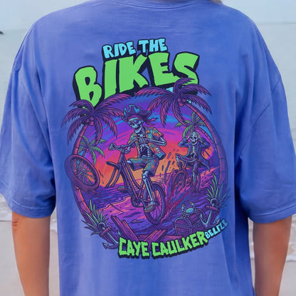 Person wearing a blue t-shirt with 'Ride the Bikes' Pirate skeleton cartoon graphic on a beach