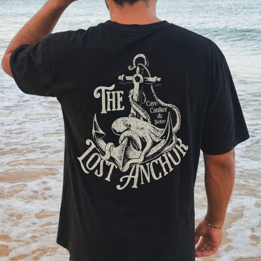 Man standing on a beach wearing a black t-shirt with 'The Lost Anchor' design.